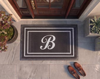 May include: A rectangular doormat featuring a dark gray textured surface with a white double-line border and a large white script letter B centered in the middle. The mat is positioned on a light gray tiled floor in front of a wooden door.