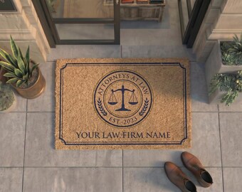May include: A rectangular tan coir doormat featuring a dark blue border and a central circular emblem with a scale of justice icon. The text reads ATTORNEYS AT LAW, EST. 2023, and YOUR LAW FIRM NAME in a dark blue serif font centered on the mat.