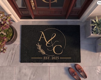 May include: A rectangular black doormat featuring a gold-colored monogram with the letters A and C, a circular line, a leaf branch graphic, and the text EST. 2025 centered horizontally below the monogram.