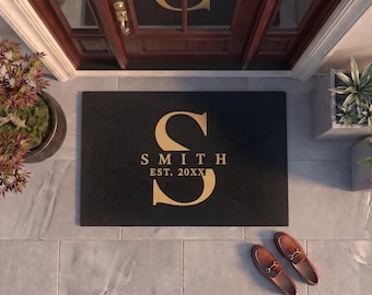 May include: A rectangular black doormat featuring a large gold-colored letter S in the center. Below the letter, the text SMITH is printed in gold, followed by the text EST. 20XX in a smaller gold font. The mat has a clean, flat surface.
