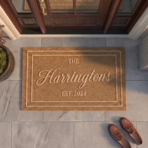 May include: A rectangular tan coir doormat featuring a double-line border and centered text that reads THE Harringtons EST. 2024 in a mix of serif and script fonts. The mat is positioned on a light gray tiled floor in front of a dark wood door.