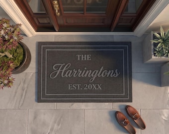May include: A rectangular dark gray doormat featuring a double-line border and centered text that reads THE Harringtons EST. 20XX in a light gray font. The mat is positioned on a light gray tiled floor in front of a dark wood door.