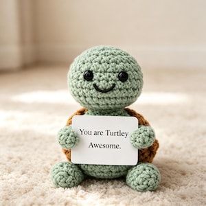 May include: A light green crocheted turtle plush toy with a brown shell, black plastic eyes, and a stitched smile. The turtle holds a white rectangular card featuring the black printed text, You are Turtley Awesome. The toy has a soft texture.