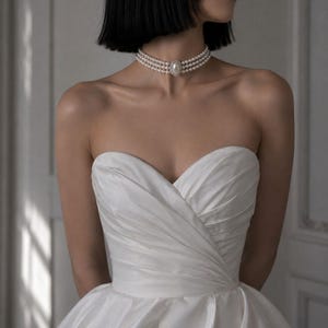 Puede incluir: Vestido de novia blanco de satén sin tirantes, con escote corazón y falda amplia. El corpiño es entallado. Se lleva un collar de perlas de tres hilos con un adorno central ovalado. La persona tiene el pelo corto y negro.