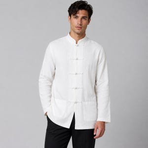 May include: A long-sleeved white shirt featuring a mandarin collar and a front closure with five fabric-covered toggle buttons. The shirt is crafted from a textured fabric and includes two large patch pockets at the lower front hem area.