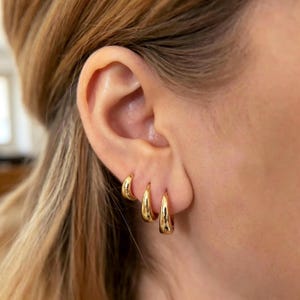 Puede incluir: Tres pendientes de aro dorados de diferentes tamaños se lucen en una oreja. Los pendientes son brillantes y reflejan la luz. La oreja está situada a la derecha, con el pelo de la persona visible a la izquierda.