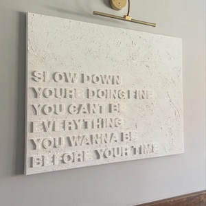 May include: A rectangular white canvas wall art piece featuring a heavily textured, impasto-style surface. The artwork displays raised, three-dimensional white lettering that reads: SLOW DOWN YOU'RE DOING FINE YOU CAN'T BE EVERYTHING YOU WANNA BE BEFORE YOUR TIME.