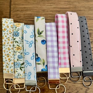 May include: A collection of fabric key fobs with gold or silver hardware. The patterns include floral, bow, blueberry, gingham, polka dot, and solid gray designs. Each fob has a metal ring for attaching keys.