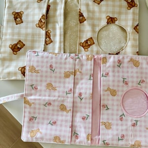 May include: Two rectangular fabric organizers with fold-over flaps. The top organizer features a beige gingham pattern with brown teddy bear illustrations. The bottom organizer has a pink gingham pattern with rabbits and small pink flowers.