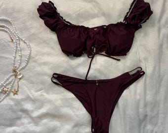 May include: A dark burgundy two-piece bikini set featuring a ruffled off-the-shoulder top with a center lace-up tie detail and matching bikini bottoms with side strap accents. The set is displayed alongside a white pearl necklace and a starfish.