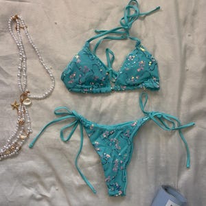 May include: A turquoise two-piece bikini set featuring a triangle top with halter ties and matching side-tie bottoms, both adorned with scattered iridescent sequins. A long white pearl necklace with small seashell charms and a blue candle are nearby.