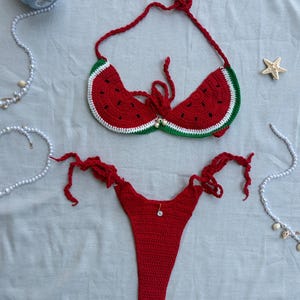 Puede incluir: Un conjunto de bikini de ganchillo de dos piezas con diseño de sandía. La parte superior tiene copas rojas con detalles de semillas negras, borde blanco y verde con tiras para atar. La parte inferior roja a juego tiene lazos laterales y un dije.