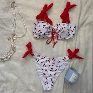 May include: A two-piece bikini set featuring a white fabric with a red cherry print. The top has red tie-shoulder straps and a center front tie. The bikini bottoms feature red tie-side closures. A pearl necklace with small seashells and a light blue candle.
