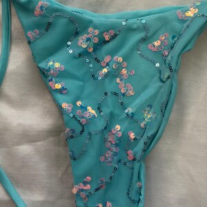 May include: A turquoise bikini bottom crafted from a textured mesh fabric. The garment is adorned with scattered, iridescent, circular sequins in shades of pink, blue, and gold, arranged in swirling patterns across the front surface.