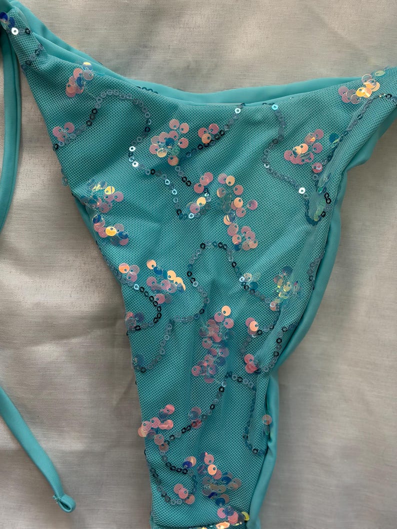 May include: A turquoise bikini bottom crafted from a textured mesh fabric. The garment is adorned with scattered, iridescent, circular sequins in shades of pink, blue, and gold, arranged in swirling patterns across the front surface.