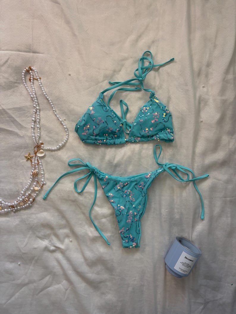 May include: A turquoise two-piece bikini set featuring a triangle top with halter ties and matching side-tie bottoms, both adorned with scattered iridescent sequins. A long white pearl necklace with small seashell charms and a blue candle are nearby.
