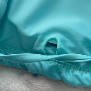 May include: A bright turquoise fabric garment featuring a small, reinforced circular opening or loop stitched into the center of the material. The fabric has a smooth, slightly reflective texture with visible horizontal stitching along the bottom edge.