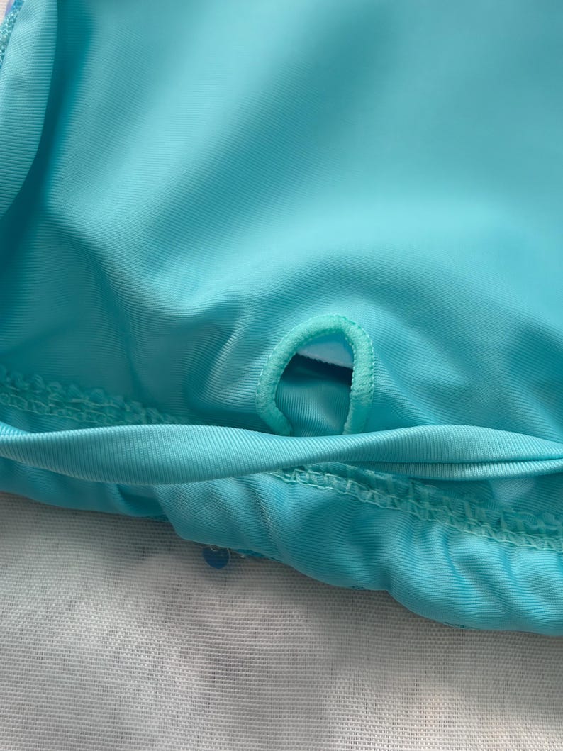 May include: A bright turquoise fabric garment featuring a small, reinforced circular opening or loop stitched into the center of the material. The fabric has a smooth, slightly reflective texture with visible horizontal stitching along the bottom edge.
