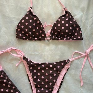 May include: A two-piece bikini set featuring a dark brown fabric with a light pink polka dot pattern. The triangle top has light pink halter straps and small bows at the top of each cup. The matching bikini bottom has side ties in light pink.