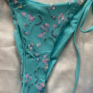 May include: A turquoise bikini bottom featuring a mesh overlay adorned with iridescent pink and blue sequins arranged in swirling patterns. The garment includes side tie closures and solid turquoise fabric panels along the edges for a secure fit.