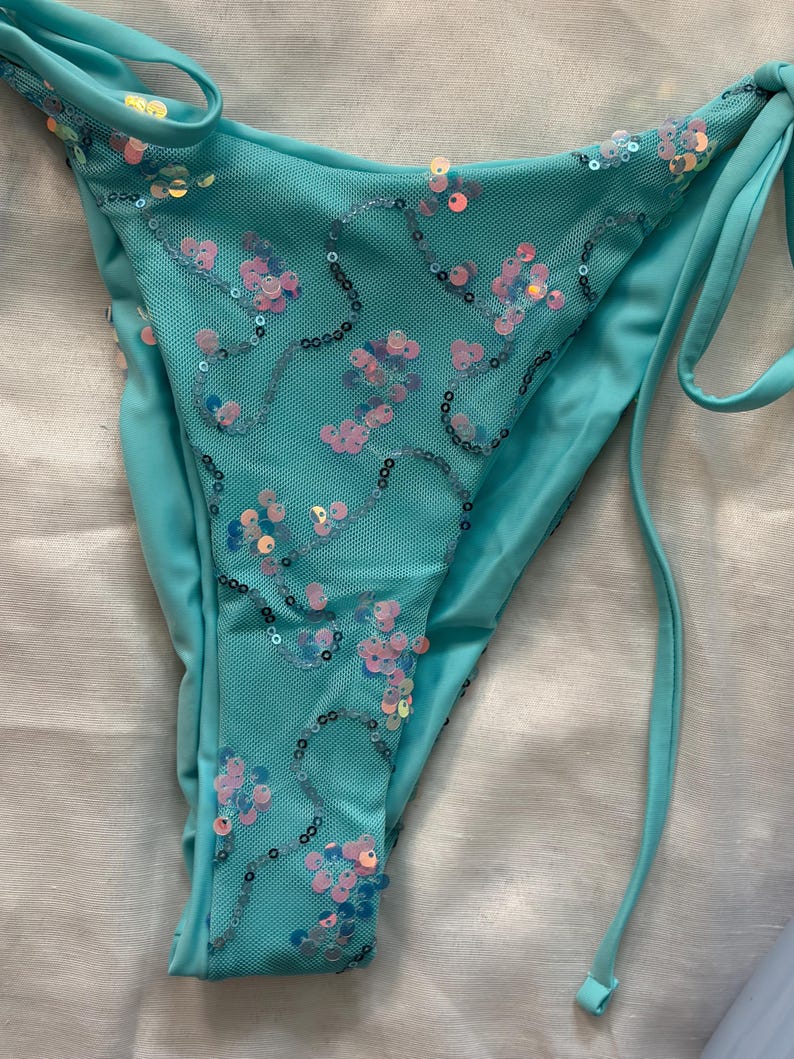 May include: A turquoise bikini bottom featuring a mesh overlay adorned with iridescent pink and blue sequins arranged in swirling patterns. The garment includes side tie closures and solid turquoise fabric panels along the edges for a secure fit.