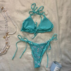 May include: A turquoise two-piece bikini set featuring a triangle top with tie closures and matching bikini bottoms with side ties. The bikini bottoms are embellished with small, shimmering pink sequins arranged in a delicate floral pattern.