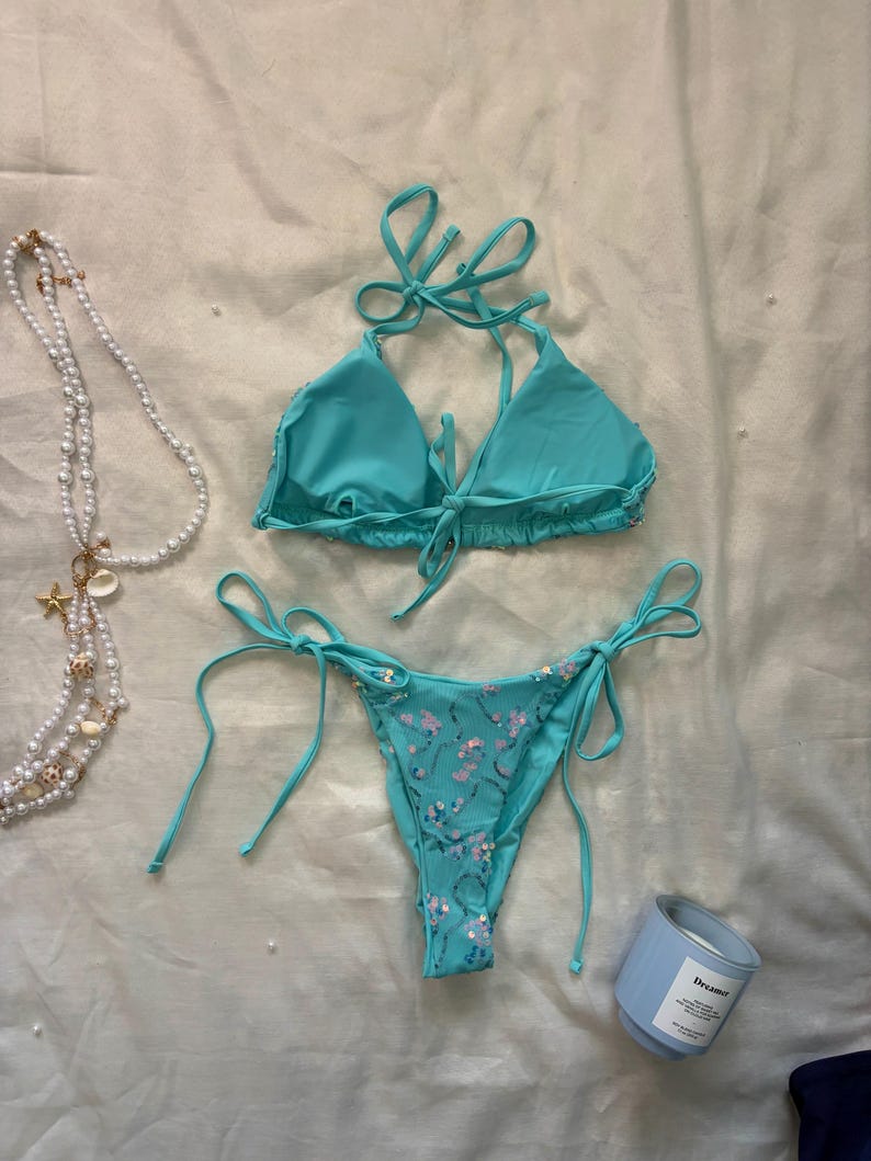 May include: A turquoise two-piece bikini set featuring a triangle top with tie closures and matching bikini bottoms with side ties. The bikini bottoms are embellished with small, shimmering pink sequins arranged in a delicate floral pattern.
