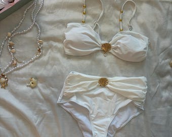 May include: A white two-piece bikini set featuring a bandeau top with gold-toned shell accents and gold beads on the straps, paired with high-waisted bottoms featuring a central gold shell detail. A long pearl necklace with small seashells is included.