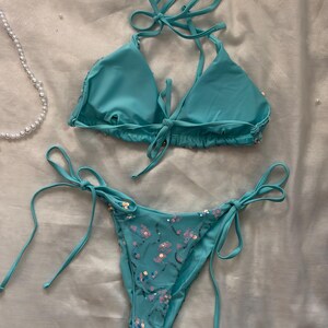 May include: A turquoise two-piece bikini set featuring a triangle top with halter neck ties and a matching bottom with side ties. The bikini is embellished with scattered iridescent sequins arranged in a swirling pattern across the fabric.