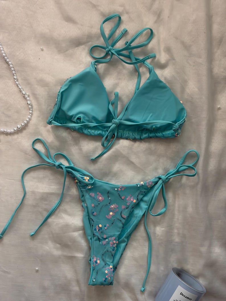 May include: A turquoise two-piece bikini set featuring a triangle top with halter neck ties and a matching bottom with side ties. The bikini is embellished with scattered iridescent sequins arranged in a swirling pattern across the fabric.