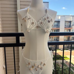 Puede incluir: Un conjunto de bikini de ganchillo blanco que incluye un top halter y una braga a juego. La prenda está adornada con varias conchas pequeñas de color natural fijadas a la tela. Los tirantes del top incluyen cuentas de madera y la braga se ata en los laterales.
