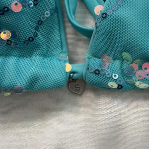May include: A turquoise bikini top crafted from mesh fabric, adorned with iridescent sequins in circular patterns. A small, silver-toned heart-shaped charm featuring the letter S is attached to the center bridge between the two cups.