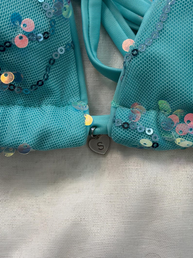 May include: A turquoise bikini top crafted from mesh fabric, adorned with iridescent sequins in circular patterns. A small, silver-toned heart-shaped charm featuring the letter S is attached to the center bridge between the two cups.