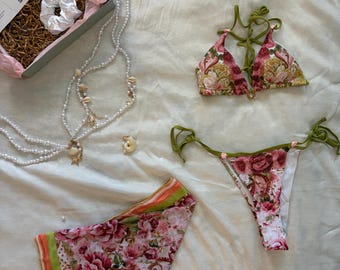 May include: A floral bikini set with olive green ties, a matching floral scarf with orange and green borders, a multi-strand pearl necklace with seashell accents, a white satin scrunchie, and a small seashell charm, all presented in a gift box.