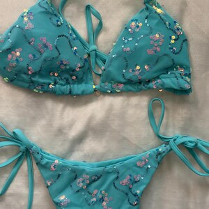 May include: A turquoise two-piece bikini set featuring a triangle top and tie-side bottoms. The fabric is covered in a mesh overlay adorned with iridescent, multi-colored sequins arranged in swirling patterns. The top includes thin halter straps.