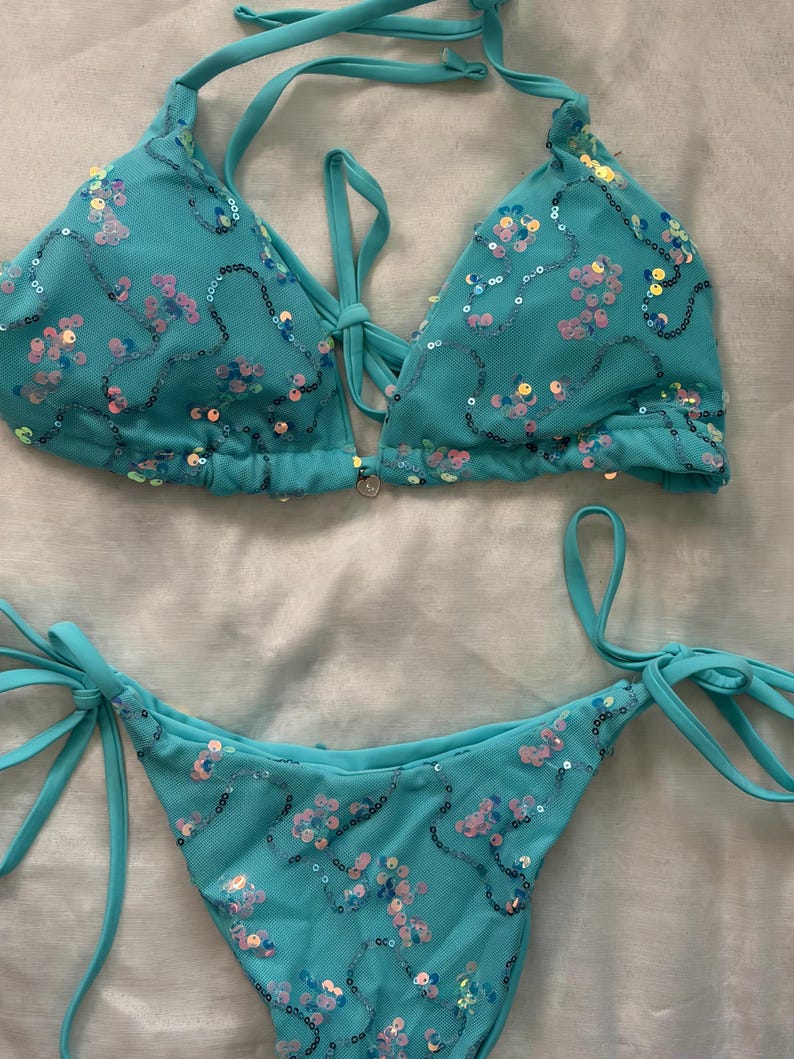 May include: A turquoise two-piece bikini set featuring a triangle top and tie-side bottoms. The fabric is covered in a mesh overlay adorned with iridescent, multi-colored sequins arranged in swirling patterns. The top includes thin halter straps.