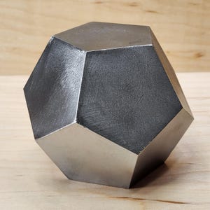 Pode incluir: Uma escultura geométrica metálica multifacetada com um acabamento escovado. O objeto é um poliedro, com uma cor cinza escuro em algumas faces e uma cor prateada mais clara em outras. A escultura é exibida em uma superfície de madeira clara.