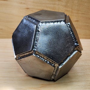 Könnte beinhalten: Eine geometrische Metallskulptur mit einem vielschichtigen Design. Die Skulptur besteht aus geschweißten Metallteilen, wodurch eine einzigartige, industrielle Ästhetik entsteht. Das silberfarbene Objekt wird auf einer Holzoberfläche präsentiert.
