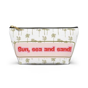 Puede incluir: Bolsa de cosméticos blanca con cremallera dorada y un estampado repetido de palmeras. La bolsa presenta el texto "Sun, sea and sand!" en rosa y rojo.