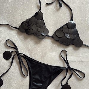 May include: A black bikini set featuring a triangle top adorned with overlapping circular black sequins. The matching bikini bottom has side ties finished with dangling circular black sequin accents, creating a cohesive and textured appearance.