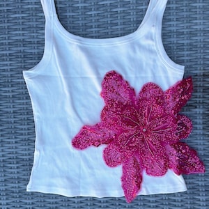 May include: A white ribbed vest top featuring a large, vibrant hot pink floral appliqué on the front. The flower is heavily embellished with matching pink sequins that cover the petals, creating a textured and reflective surface across the design.