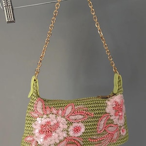Op de afbeelding: Een kleine olijfgroene geweven handtas met een bloemmotief van roze kralen en pailletten. De tas heeft een goudkleurige metalen kettinghengsel en lichtgroene zijlipjes, met een ritssluiting langs de bovenrand om persoonlijke spullen op te bergen.