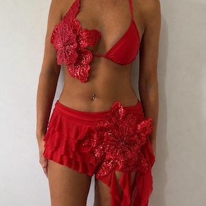 Puede incluir: Un conjunto de dos piezas rojo que consta de un top de bikini estilo halter y una falda con volantes. Ambas piezas están adornadas con grandes aplicaciones florales rojas tridimensionales cubiertas de lentejuelas brillantes.