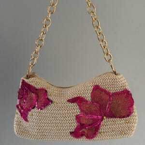 May include: A beige woven handbag featuring two large, embroidered fuchsia floral appliques with sequin accents. The bag has a gold-tone metal chain strap and small gold-tone studs scattered across the front surface of the textured material.