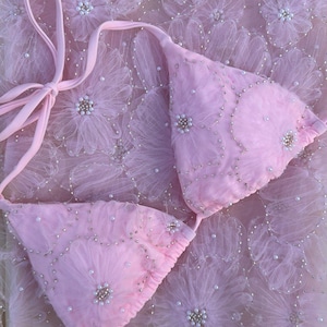 May include: A light pink bikini top featuring triangular cups adorned with layered floral fabric appliques, small white pearls, and silver bead accents. The garment includes thin matching pink fabric ties for closure and adjustment.
