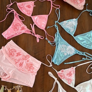 Op de afbeelding: Een collectie van drie kralenhandtassen en drie bikinysets in roze, lichtblauw en wit. De handtassen zijn voorzien van bloemvormig kralenwerk en kettinghengsels. De bikinysets bevatten driehoekige tops en broekjes met bijpassende bloemversieringen.