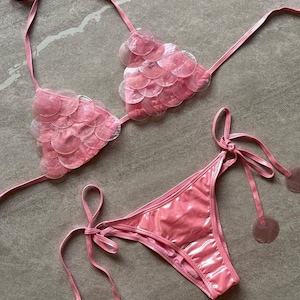 May include: A pink bikini set featuring a triangle top and matching bottoms. The top is adorned with overlapping translucent circular discs. The bottoms have side ties finished with circular disc accents. The fabric has a glossy, smooth finish.