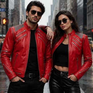 Può includere: Due persone indossano giacche di pelle rosse con dettagli grigi sulle spalle e multiple tasche con cerniera. Sono in piedi su una strada cittadina con un attraversamento pedonale ed edifici sullo sfondo. Entrambi indossano occhiali da sole.