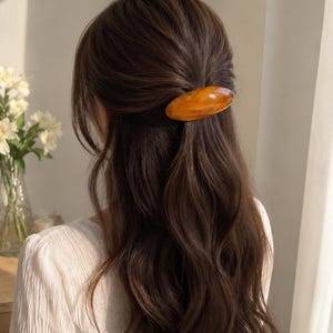 以下が含まれることがあります： 渦巻き模様が入った滑らかで光沢のある琥珀色の楕円形のヘアクリップ。このヘアアクセサリーは、長くウェーブのかかった茶色の髪に留められており、白いシャツの上で髪の一部をハーフアップスタイルにまとめています。