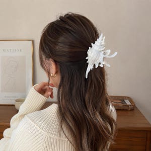 Könnte beinhalten: Eine weiße Haarspange in Form einer detaillierten Muschel. Die Spange ist in langem, dunkelbraunem Haar befestigt und hält einen Teil des Haares zurück. Das Design zeigt die texturierte, spiralförmige und spitze Struktur einer Meeresmuschel.